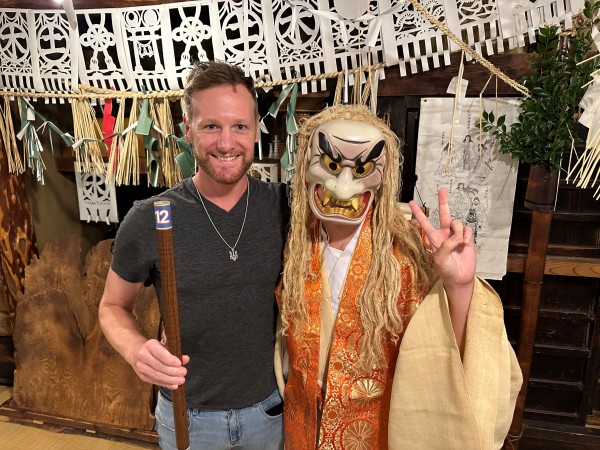 A man stands smiling with his arm around a person dressed in traditional Japanese garb, including a mask.